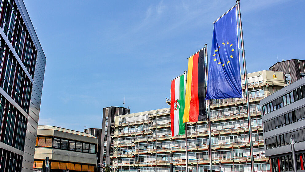 Vor dem Haupteingang der Universität Paderborn sind die Flagge des Landes Nordrhein-Westfalen, die Deutschland-Flagge und die Flagge der Europäischen Union gehisst.
